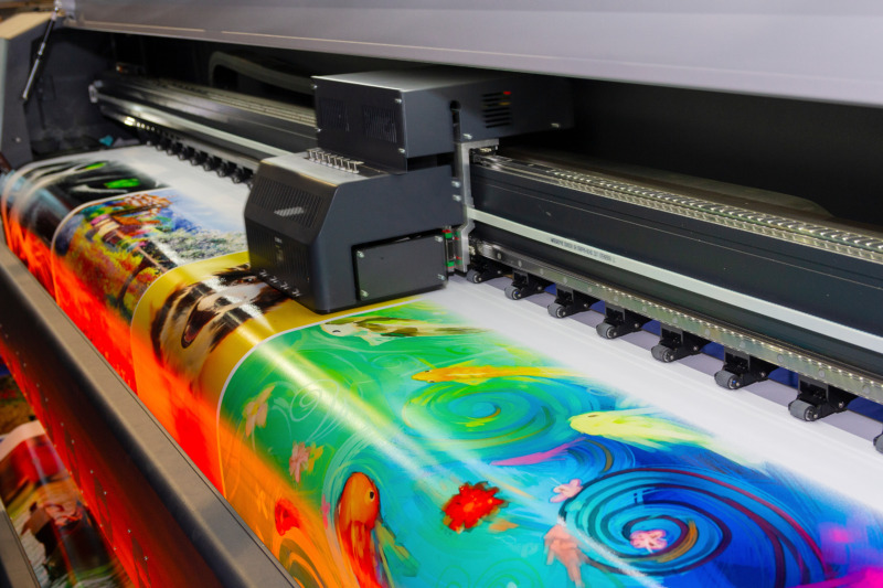 Large format printing machine in operation. Printing a large banner of brightly coloured images.