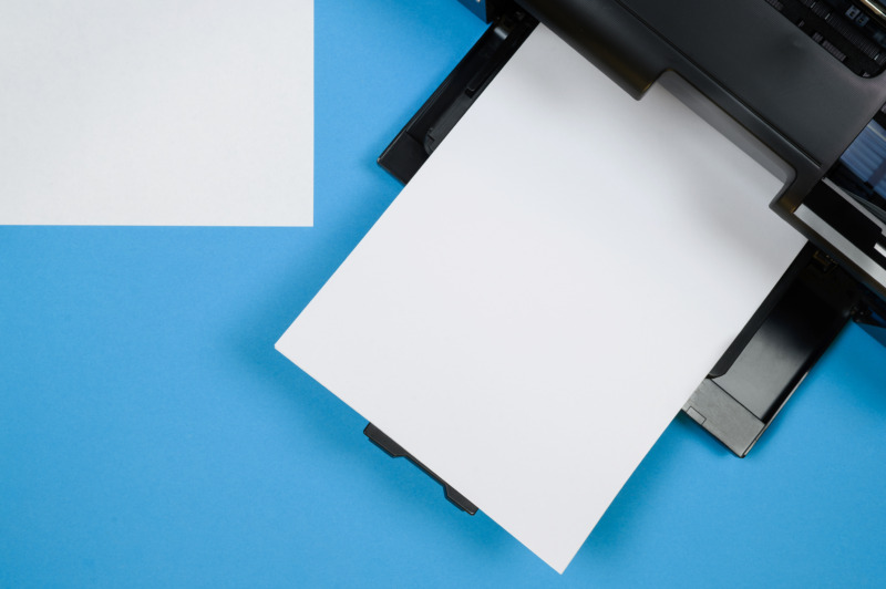 Top view of a black printer and a blank sheet of A4 paper on a blue background. Office Equipment.